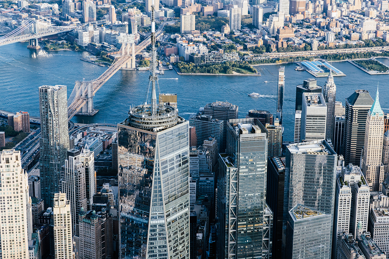 Aerial view of Lower Manhattan skyscrapers with the Brooklyn and Manhattan Bridges crossing the East River, and Brooklyn visible in the background on a clear day.