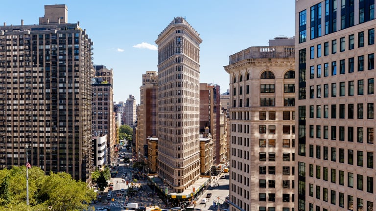 Aerial view of the Flatiron Building in New York City. The triangular building is flanked by other high-rise structures, with streets bustling with vehicles and trees visible in the foreground under a clear blue sky.