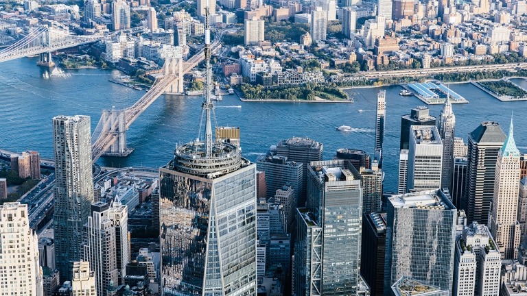 Aerial view of New York City skyscrapers with the Brooklyn Bridge and East River in the background.