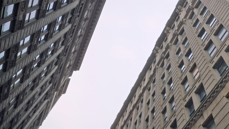  Upward view of two tall, ornate buildings in an urban setting, converging towards the sky. The buildings feature numerous windows and detailed architectural elements, set against an overcast, gray sky.