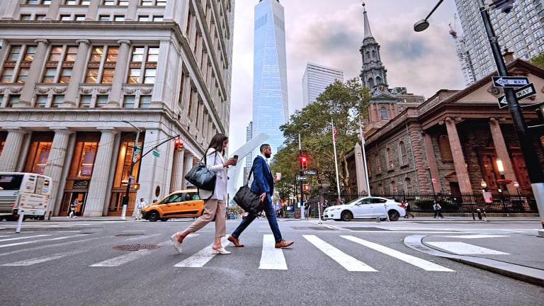 Two people cross a city street at a crosswalk, surrounded by tall buildings. The One World Trade Center tower is visible in the background. Cars, a taxi, and street signs are also present.