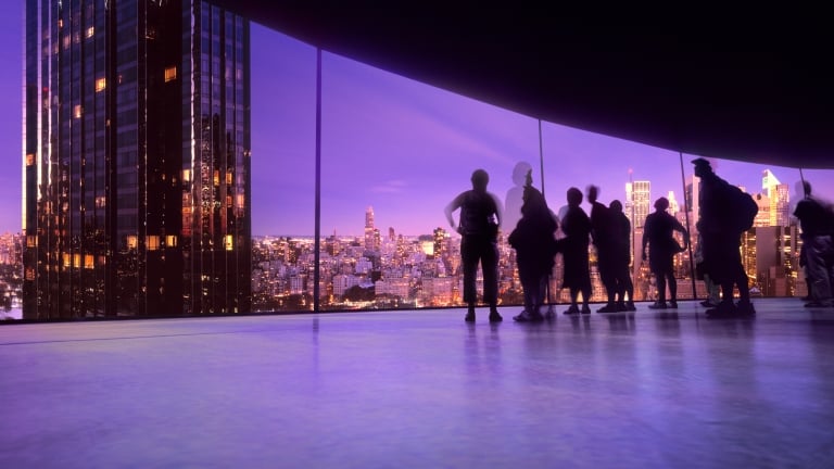 Silhouettes of people stand by large windows overlooking a cityscape at dusk, with skyscrapers and buildings illuminated under a vibrant purple sky.