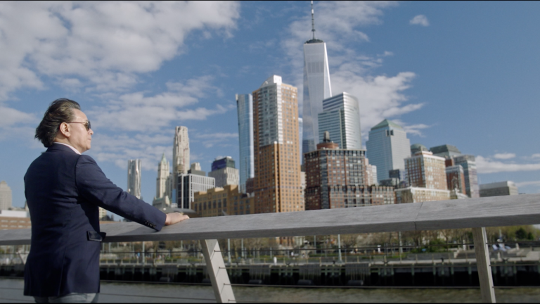Hyeon Yeo, Co-Chair of KAIST New York Campus Committee, stands in a navy blazer on a waterfront railing, looking at the New York City skyline with skyscrapers and the One World Trade Center under a partly cloudy sky.