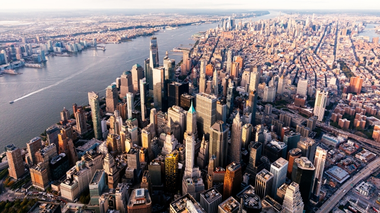 Aerial view of Manhattan, New York City, showing densely packed skyscrapers, the Hudson River on the left, and sprawling cityscape stretching into the distance under a partly cloudy sky.