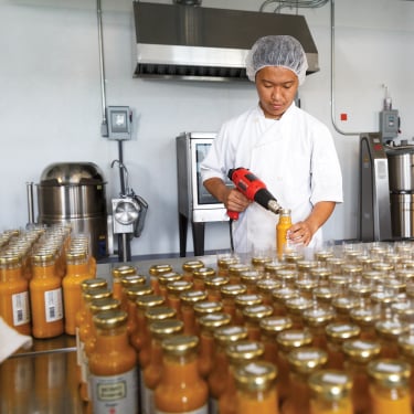 A MOMO Dressing employee in a white uniform and hairnet seals glass bottles of orange liquid with a heat gun in a food production facility. Numerous capped bottles are neatly arranged on a stainless steel table. Industrial kitchen equipment is visible in the background.