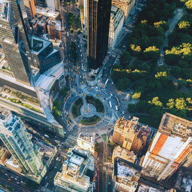 Aerial view of Columbus Circle showing a busy city intersection surrounded by tall skyscrapers, a circular plaza with a monument at the center, and a lush green park to the right.