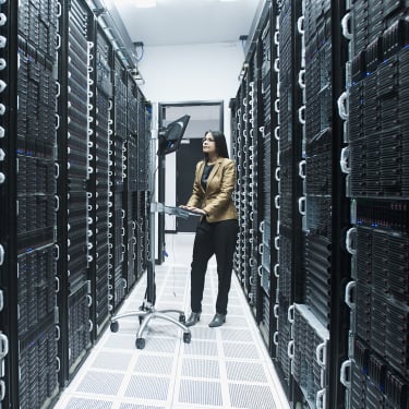 A businesswoman in a brown jacket stands in a server room, surrounded by tall racks of servers. She is working on a computer mounted on a stand. The room is well-lit with a checkered floor.