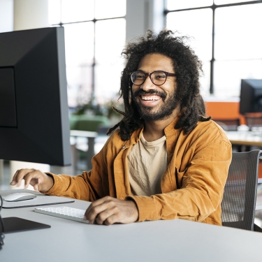 Cheerful office worker with beard, wearing casual shirt, at desk using computer.