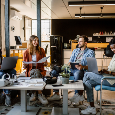 Four people sitting in a modern office, engaged in a casual meeting. They are using laptops and holding papers. The setting includes a coffee table, plants, and a kitchen area in the background, creating a relaxed atmosphere.