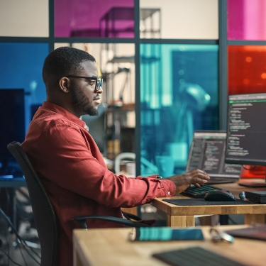 African American Man Writing Lines Of Code On Desktop Computer With Multiple Monitors and Laptop in Creative Office. African American Man Writing Lines Of Code On Desktop Computer With Multiple Monitors and Laptop in Creative Office.
