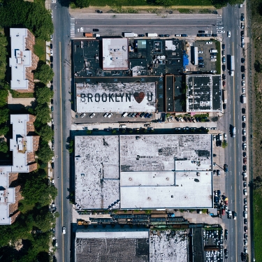 Aerial view of a city block with two large buildings. One rooftop has the text BROOKLYN ❤️ U painted on it. The block is surrounded by roads with parked cars. Green spaces and other buildings are visible to the sides.