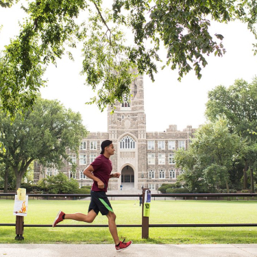 A man in athletic wear jogs on the Fordham University campus along a path lined with trees, with a large, historic stone building and green lawn in the background on a bright, clear day.