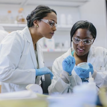 Two scientists in a lab are wearing white coats, gloves, and safety goggles. They are discussing data. Another scientist works in the background. The lab is filled with equipment and supplies.