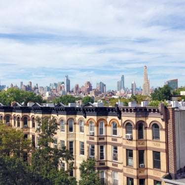 A row of tan brick apartment buildings with arched windows in the foreground, leafy trees, and a distant NYC skyline under a partly cloudy blue sky.