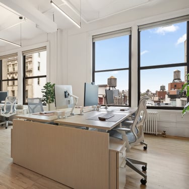 Modern, bright office space with large windows, several desks with computers, ergonomic chairs, and a view of city buildings and water towers outside. Indoor plants add greenery to the clean, minimal decor.