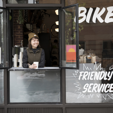 A person in a beanie smiles while holding a cup at a coffee shop window. Signs read BIKE, FRIENDLY SERVICE, and COLD BREW, with an A rating visible on the window. Indoor plants and lights are in the background.