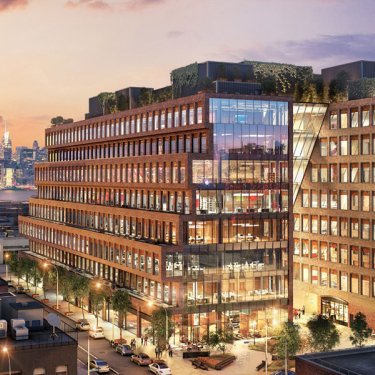 Modern office building with large glass windows and rooftop greenery, lit up at dusk with a city skyline and colorful sunset in the background. Cars and streetlights line the road in front of the building.