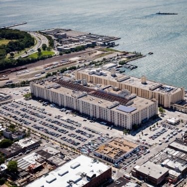 Aerial view of Brooklyn Army Terminal, a large waterfront warehouse complex with numerous parking lots filled with cars. The area is surrounded by industrial buildings, green spaces, and docks extending into the water.