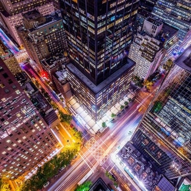 A vibrant, aerial night view of a New York City intersection surrounded by tall skyscrapers, with colorful lights from buildings and streaks of headlights and taillights showing busy street traffic below.
