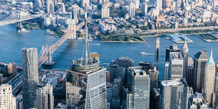 Aerial view of Lower Manhattan skyscrapers with the Brooklyn and Manhattan Bridges crossing the East River, and Brooklyn visible in the background on a clear day.