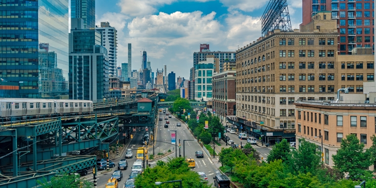 A busy urban street with cars and yellow cabs runs between tall buildings. A subway train travels on an elevated track to the left. The New York City skyline is visible in the background under a partly cloudy sky.