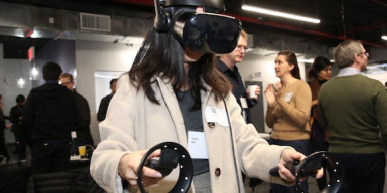 Woman wearing a tan coat uses a virtual reality headset and handheld controllers at a busy indoor event, while people in the background talk and hold drinks.