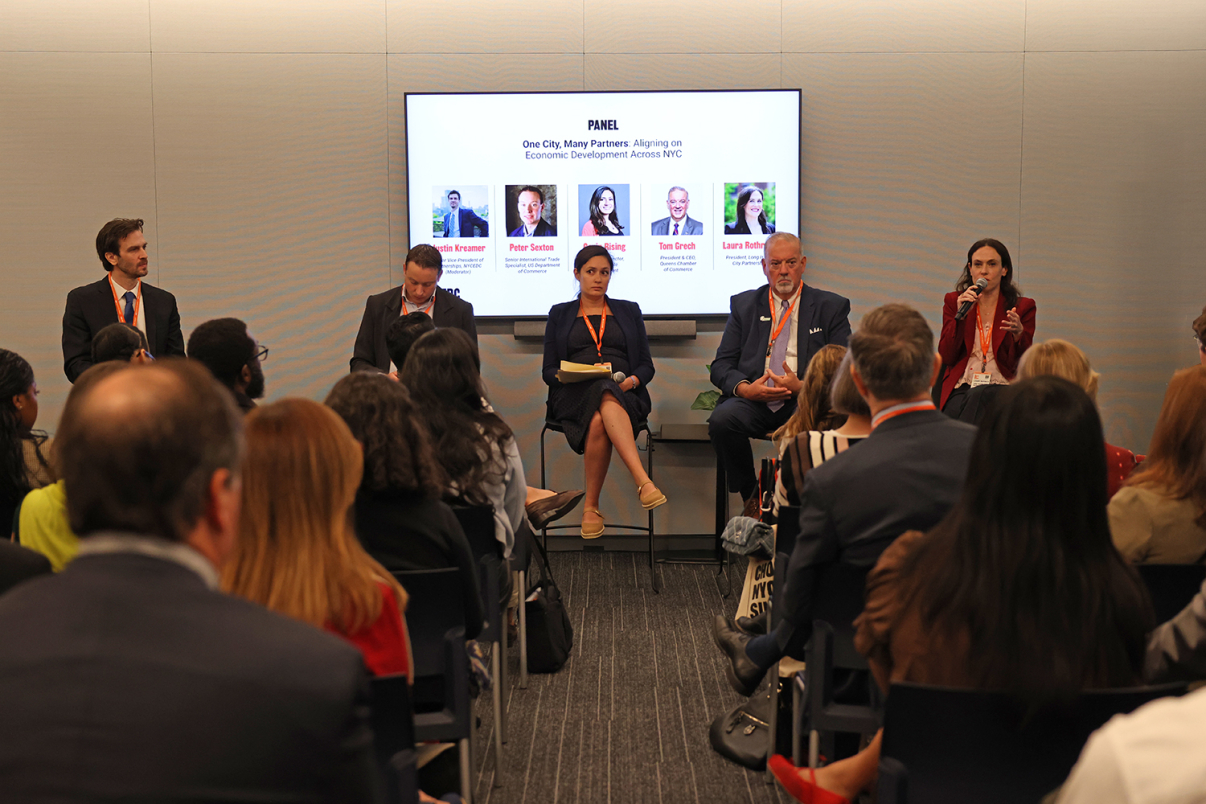A panel of five speakers sits at the front of a conference room, facing an audience. A presentation screen behind them displays their names and panel title: “One City, Many Partners: Aligning on Economic Development Across NYC.”.