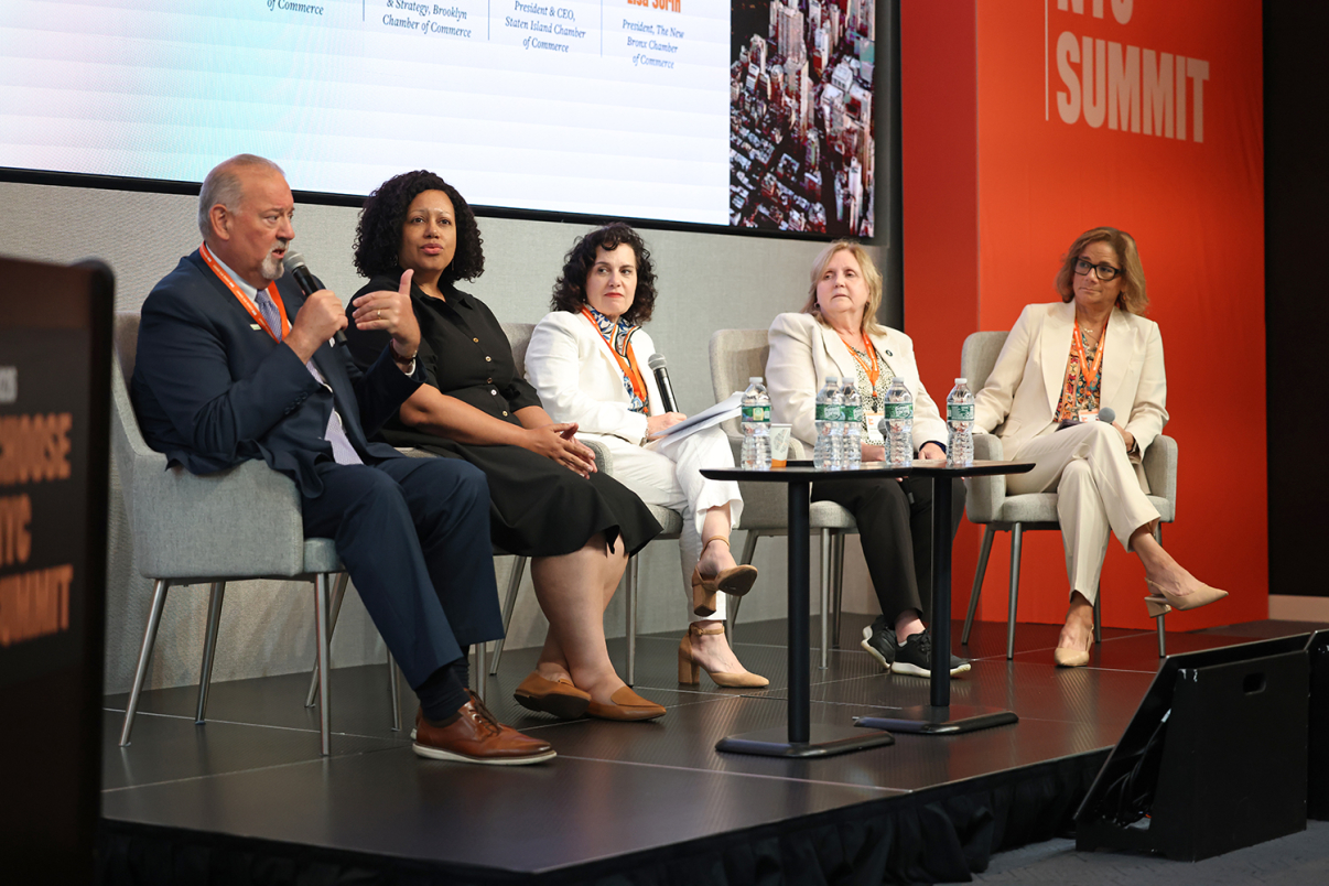 Five people in business attire sit on a stage panel, speaking and listening, with microphones and water bottles on a table. A screen and orange signs with CHOOSE NYC SUMMIT are visible in the background.