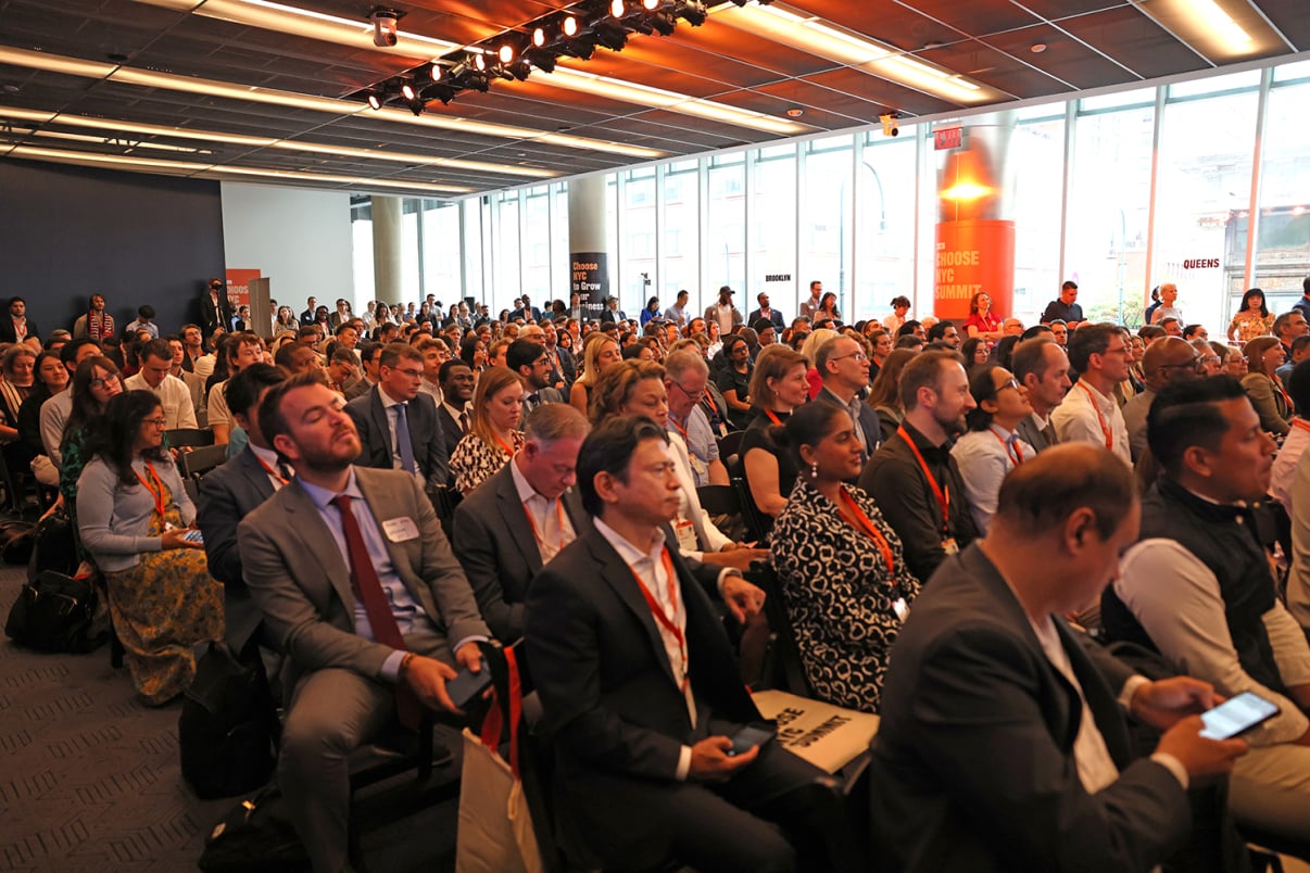 A large group of people in business attire are seated and attentively listening at a professional conference in a modern, well-lit room with large windows.