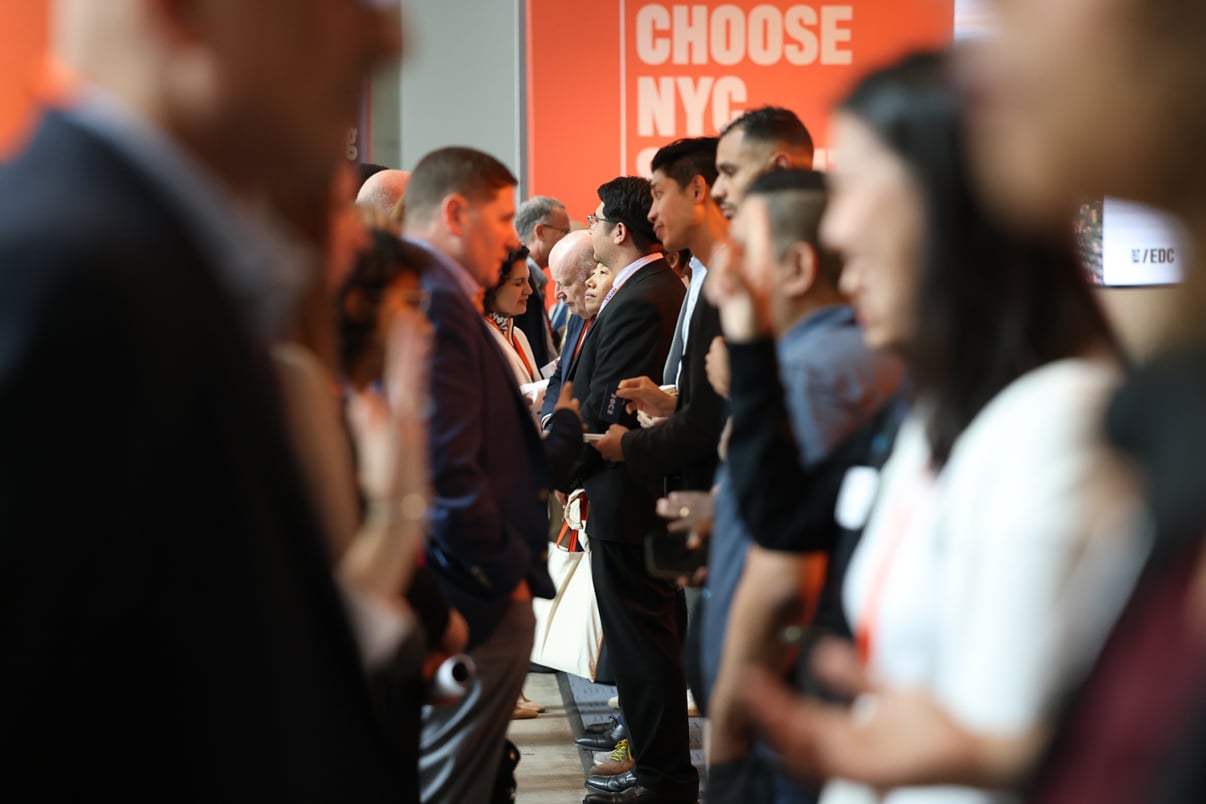 A group of people in business attire stands in two rows facing each other, engaged in conversation at a professional networking event. An orange sign in the background reads CHOOSE NYC.